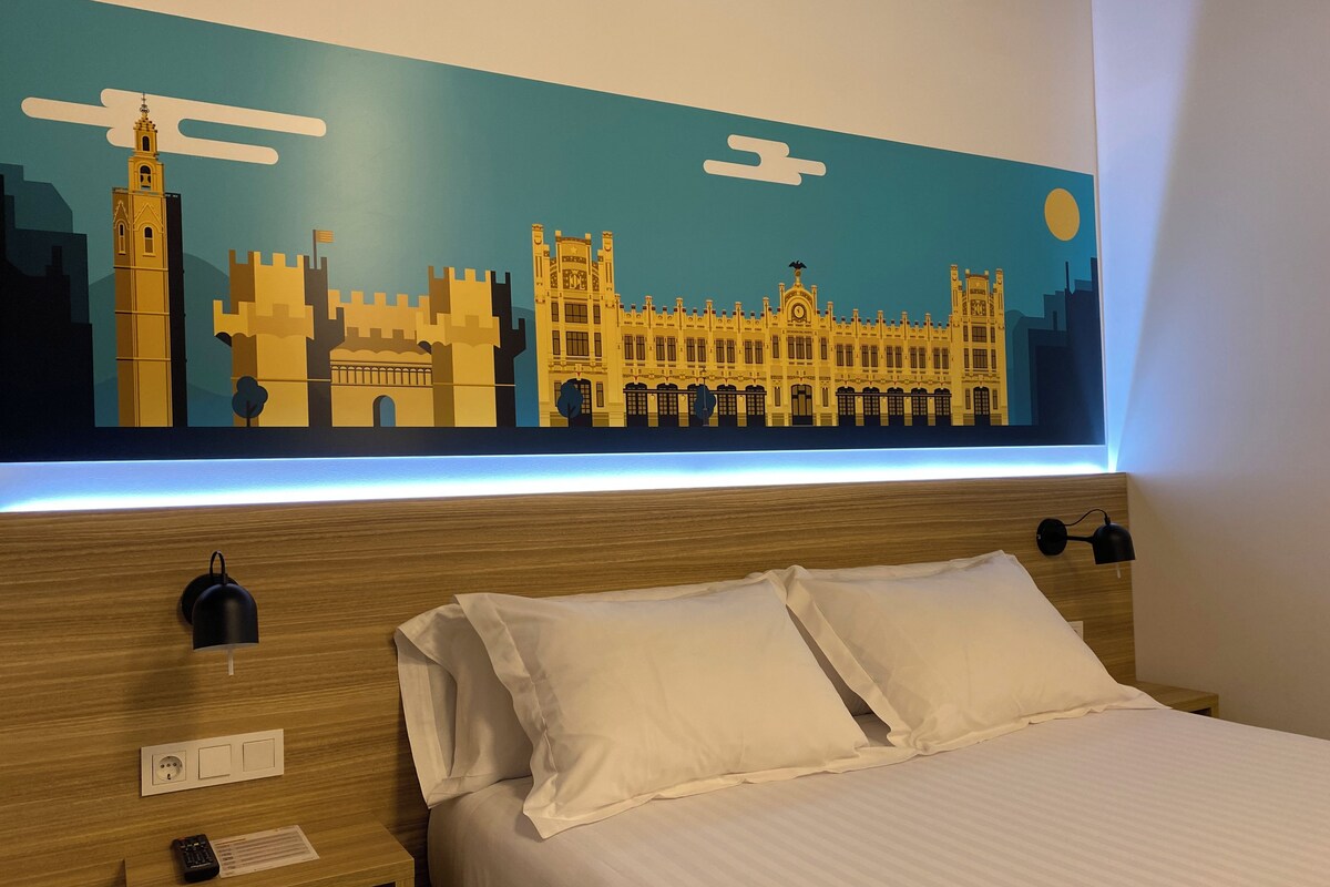 A hotel room with a bed, pillows, a nightstand, a remote control, and a wall-mounted light.