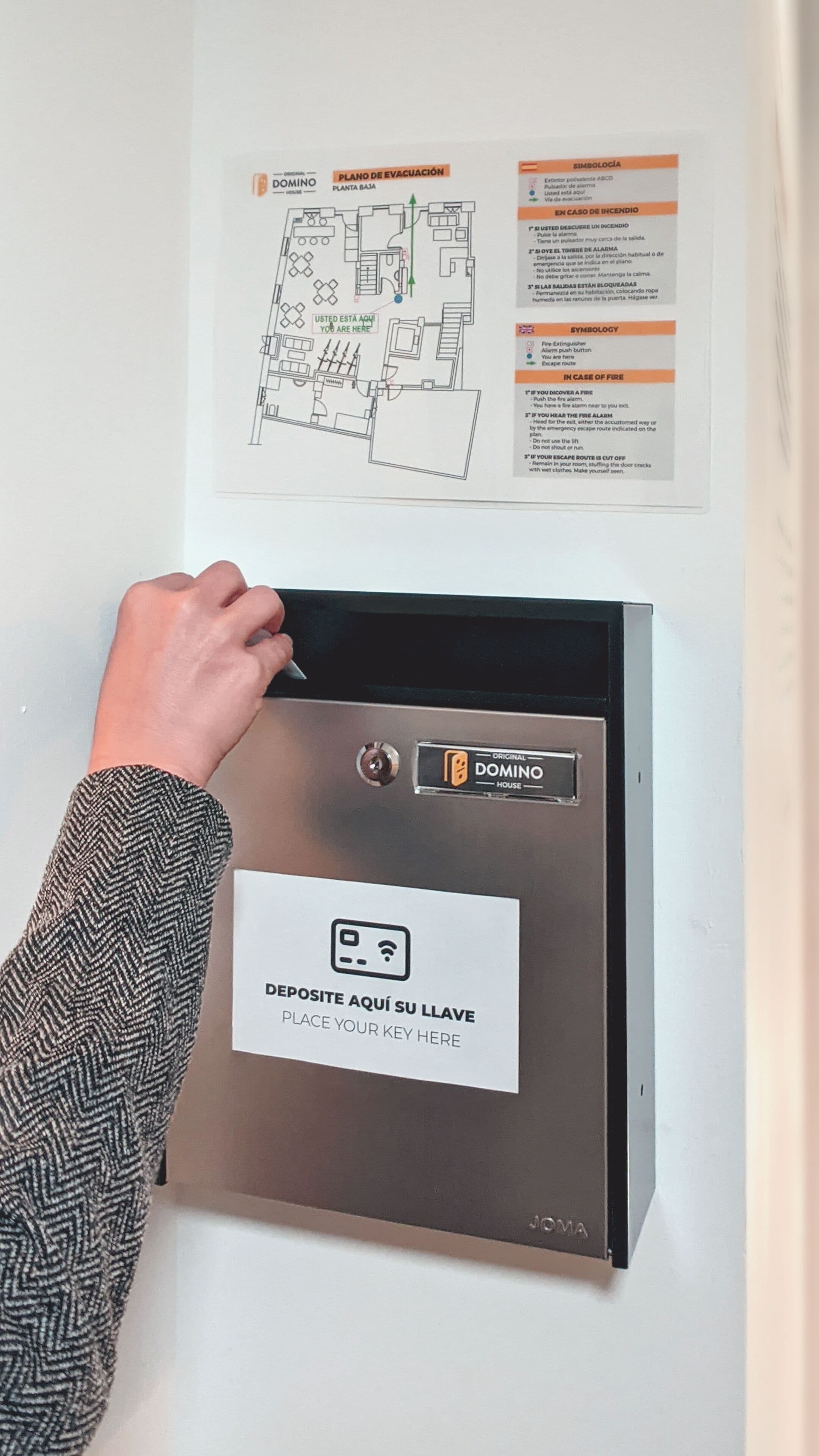 Una mano presionando un botón en un lector de tarjetas, con la leyenda 'PLACE YOUR KEY HERE' y un plano que muestra rutas de evacuación e instrucciones de emergencia.