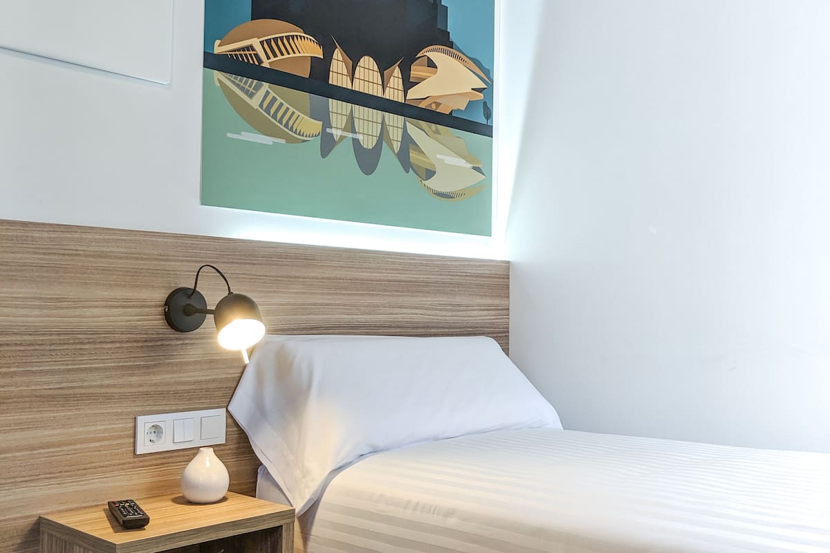 A hotel room with a bed, a nightstand with a lamp, a remote control, and a wall-mounted light switch.