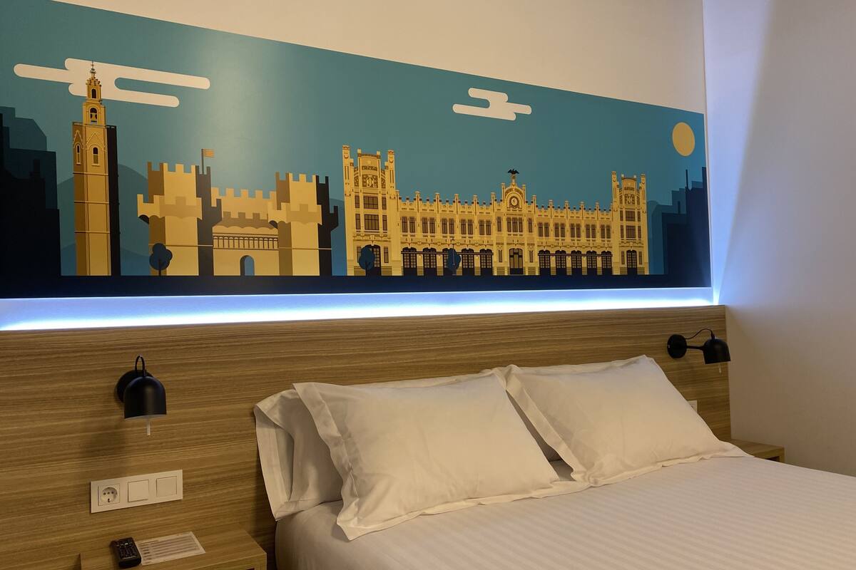 A hotel room with a bed, pillows, a nightstand, a remote control, and a wall-mounted light.