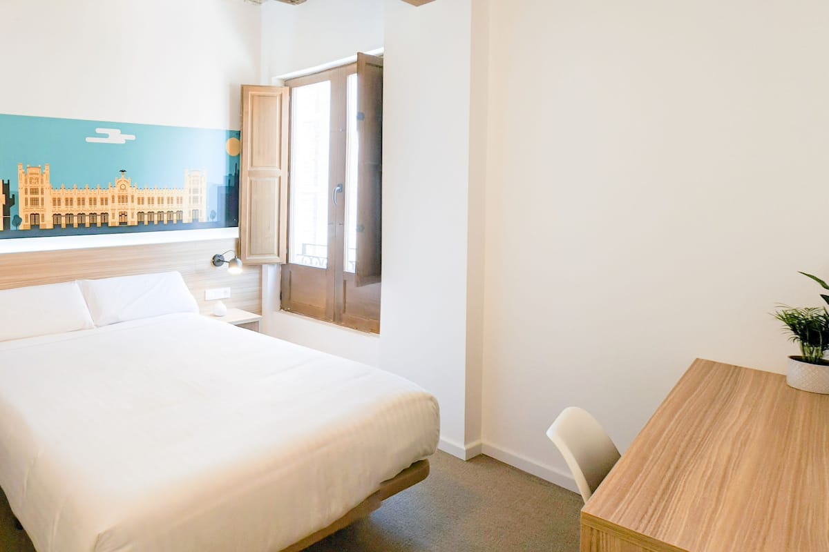 A hotel room with a bed, a nightstand, a desk, and a window with a cityscape mural.