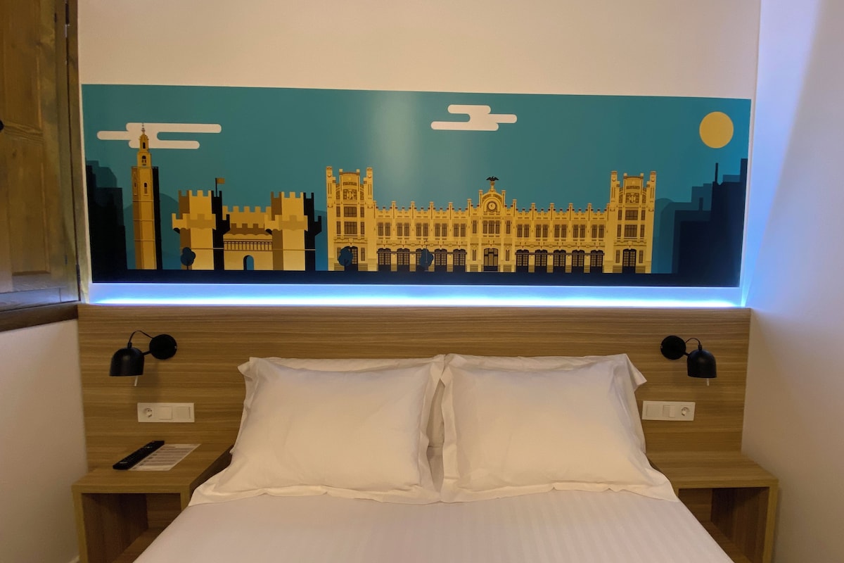 A hotel room with a bed, two bedside tables, a nightstand, a wall-mounted lamp, and a large cityscape artwork above the bed.