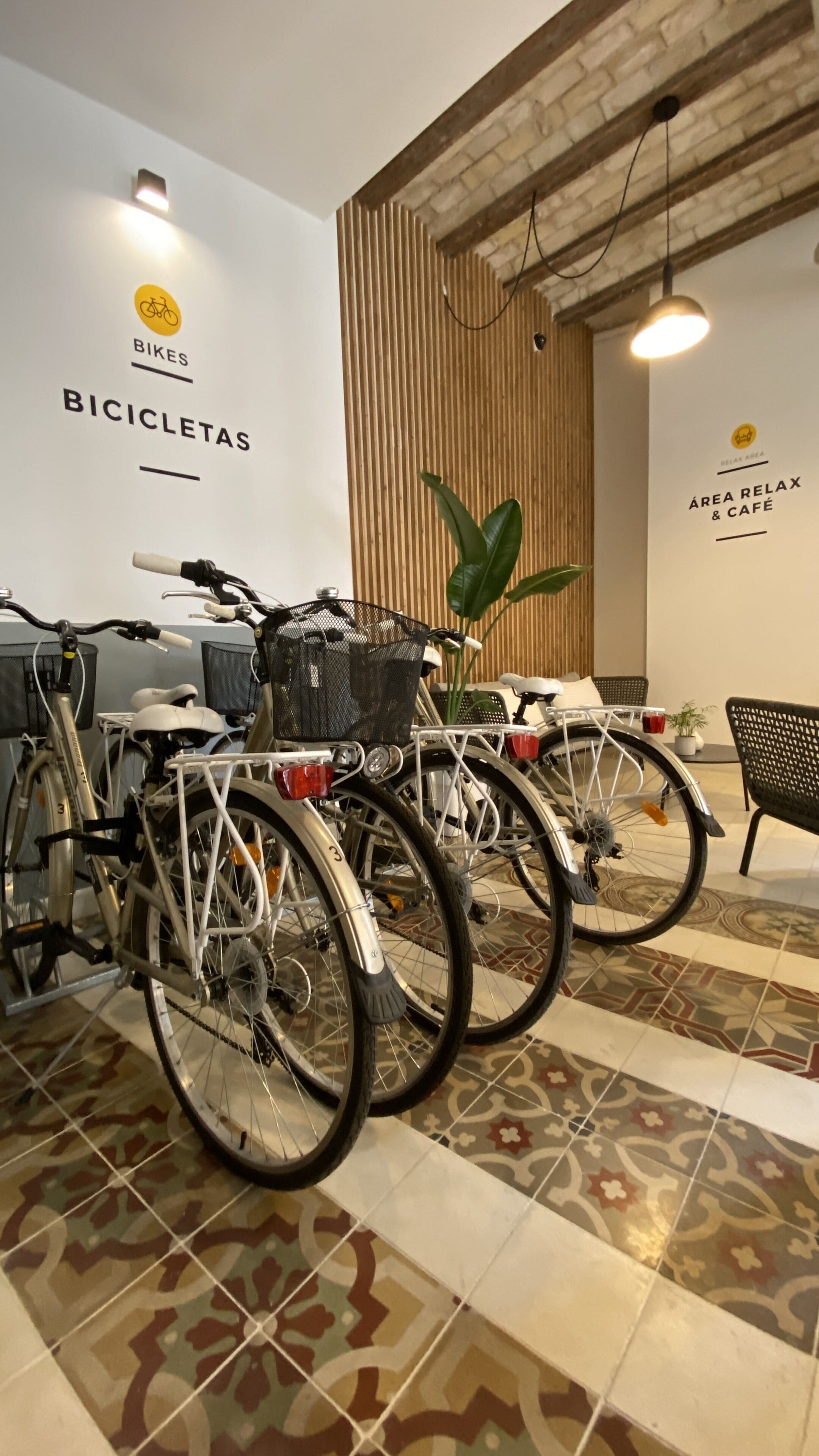 Des vélos sont garés dans une pièce dont le sol est décoré de motifs, où se trouve une plante et un panneau indiquant « BICICLETAS ».