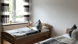 A room with two wooden beds, a window with curtains, a radiator, and a small table with a lamp. - Hostel Wendt (Fintel)