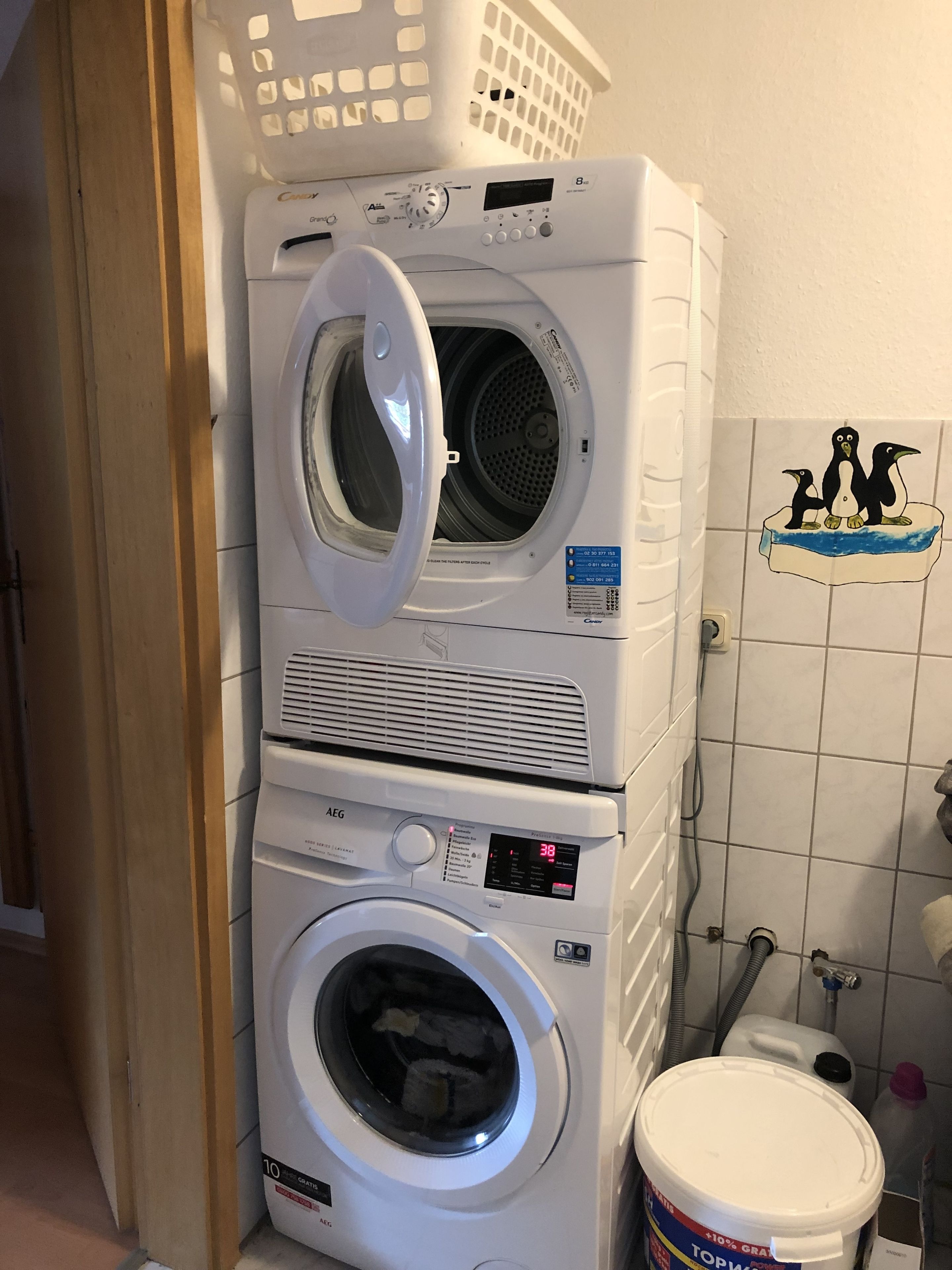 Máquina de lavar e secar empilhadas numa lavanderia com parede revestida de azulejos brancos, cesto de roupa e aquecedor de água.