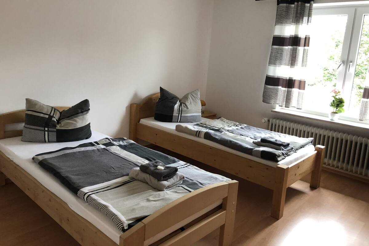 A room with two wooden beds, each with a pillow and a blanket. There is a window with curtains and a radiator below it.