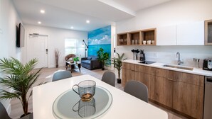 Een moderne keuken met houten kasten, een wit aanrecht en een geïntegreerde spoelbak. Er is een eethoek met een glazen tafel en stoelen, en een woonkamer met een bank en een schilderij van de oceaan.