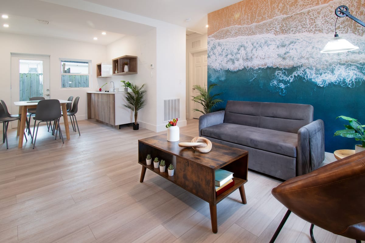 Una sala de estar moderna con un sofá gris, una mesa de centro de madera con plantas y un mural grande de olas marinas.