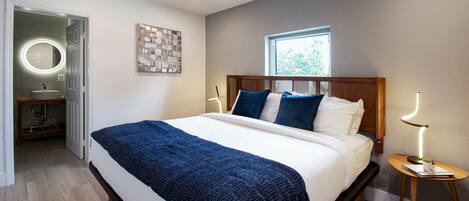 Ein Schlafzimmer mit Bett, Nachttischen, einem Fenster und einem durchgehenden Türrahmen sichtbaren Badezimmer.