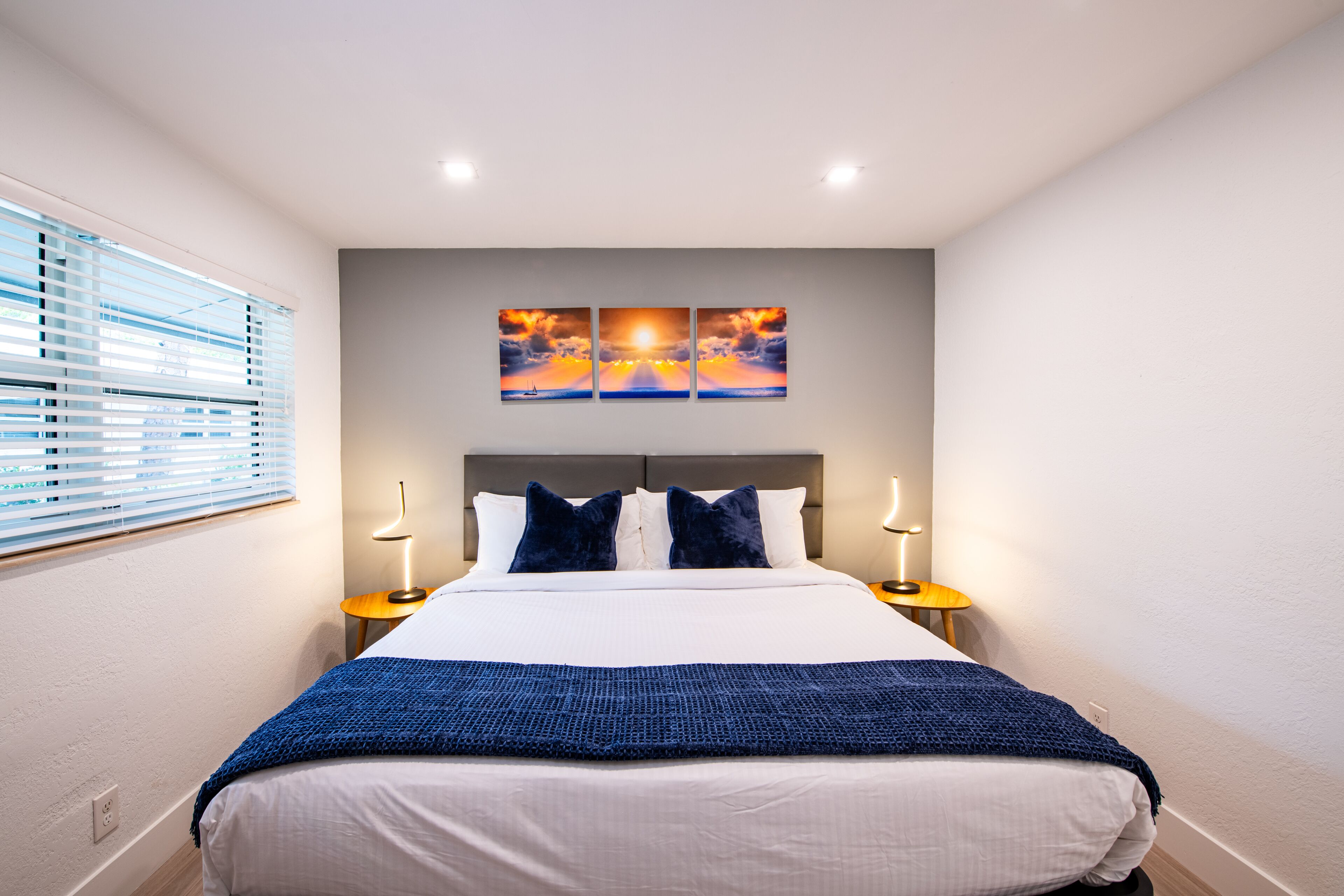A neatly made bed with a blue blanket, two bedside lamps, and a sunset painting above it.