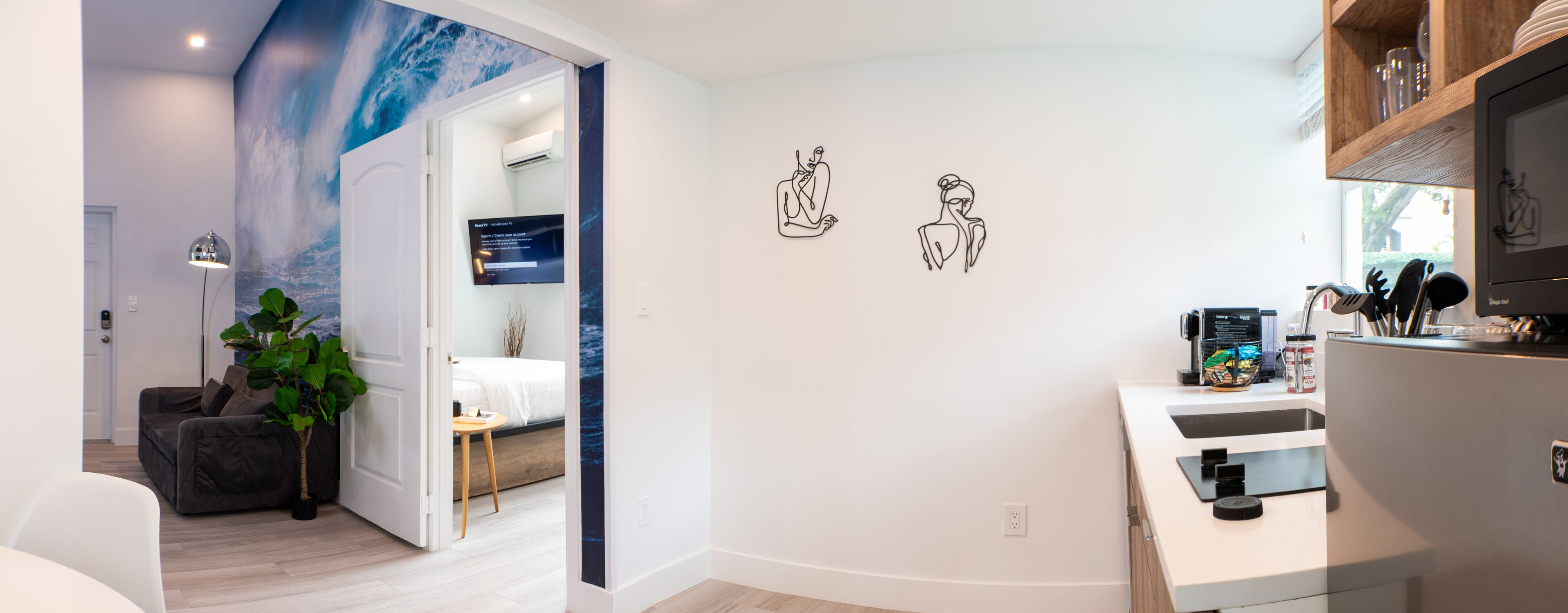 Una sala de estar moderna con un sofá gris, una mesa de centro y una planta en maceta. Se ve un dormitorio con una cama y una mesita de noche a través de una puerta abierta. La cocina incluye un refrigerador, un fregadero y diversos utensilios de cocina.