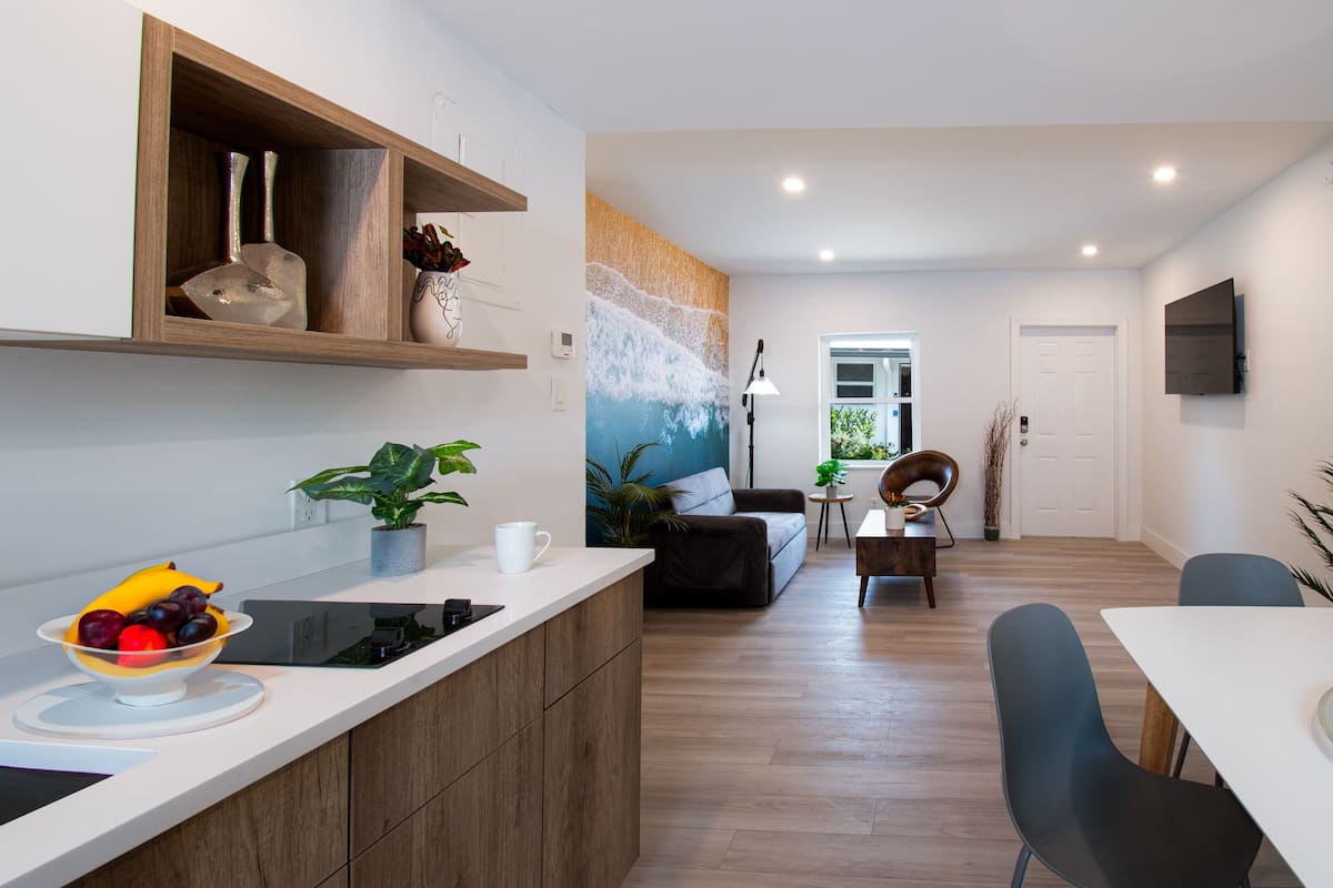Una cocina moderna con encimera de madera, un bol de fruta y una planta en maceta. Hay una zona de comedor con mesa y sillas, y una sala de estar con sofá y un cuadro grande.