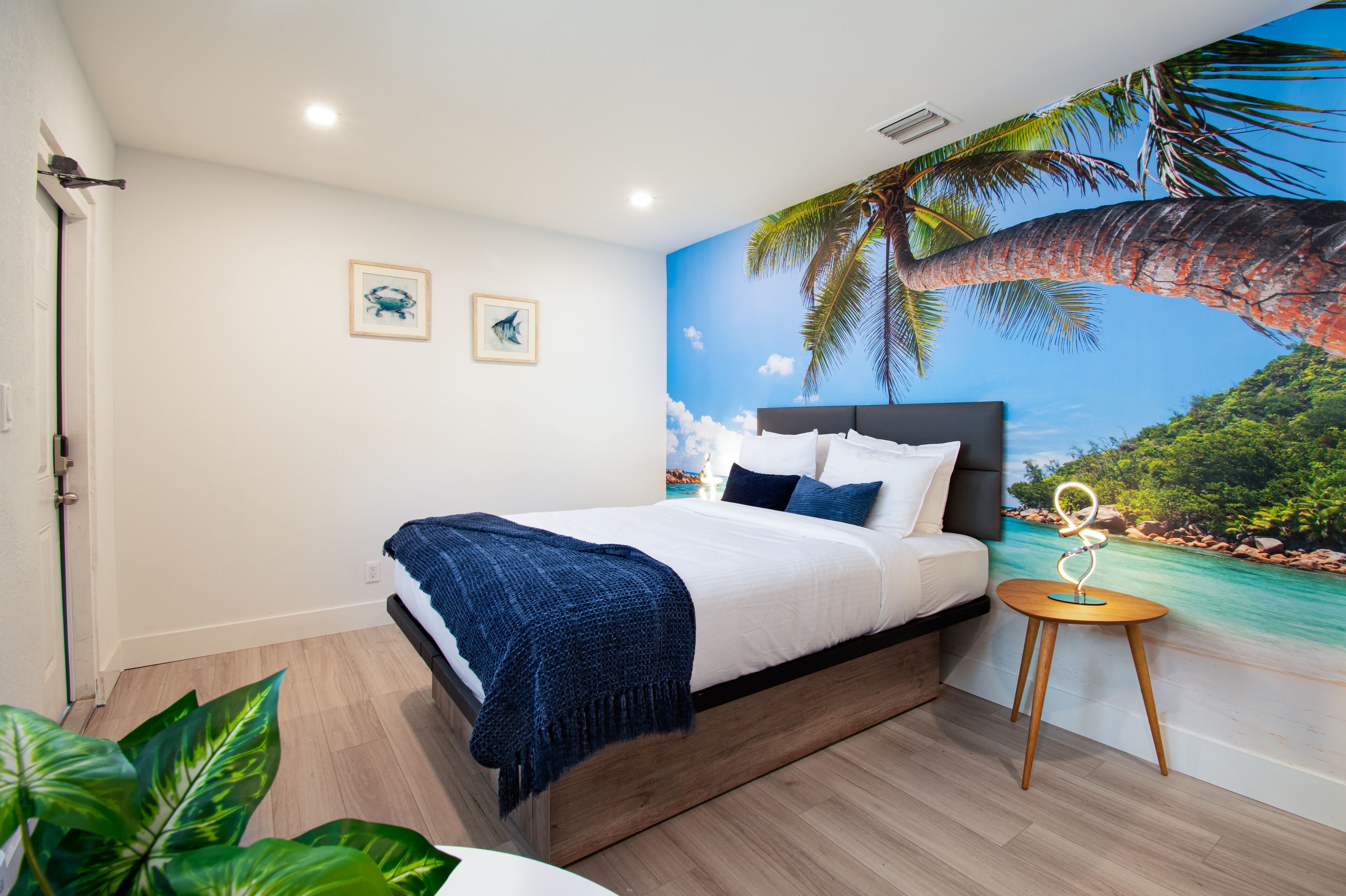 A bedroom with a bed, a nightstand, a wall mural of a beach scene, and two framed pictures on the wall.