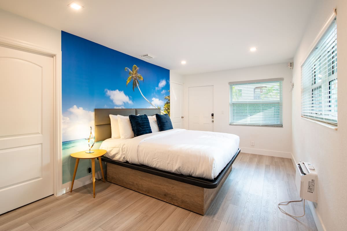 Un dormitorio con cama, mesita de noche, un mural en la pared, ventana con persianas y termostato.