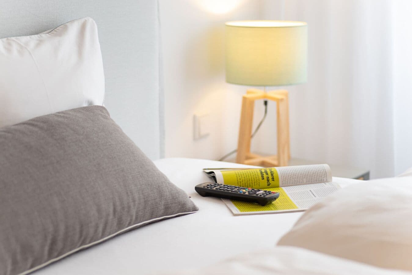 A neatly made bed with a grey pillow, a remote control, and a magazine on a white bedspread.
