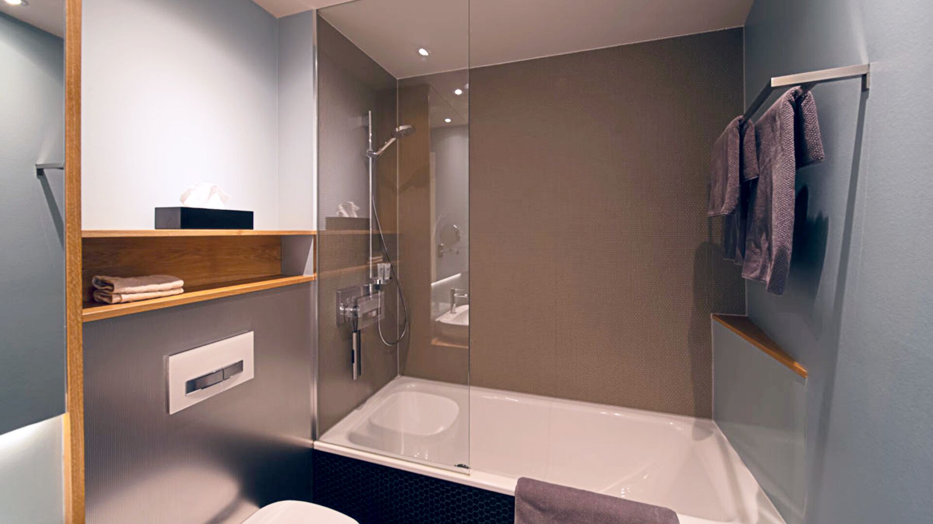 Une salle de bain moderne avec une douche à parois de verre, une baignoire et des toilettes. On y trouve des étagères en bois avec des serviettes et une petite boîte sur la gauche.