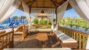 A wooden deck with a cushioned seating area, a table, and a view of a pool and palm trees. - Eftalia Ocean Resort and Spa (Alanya)