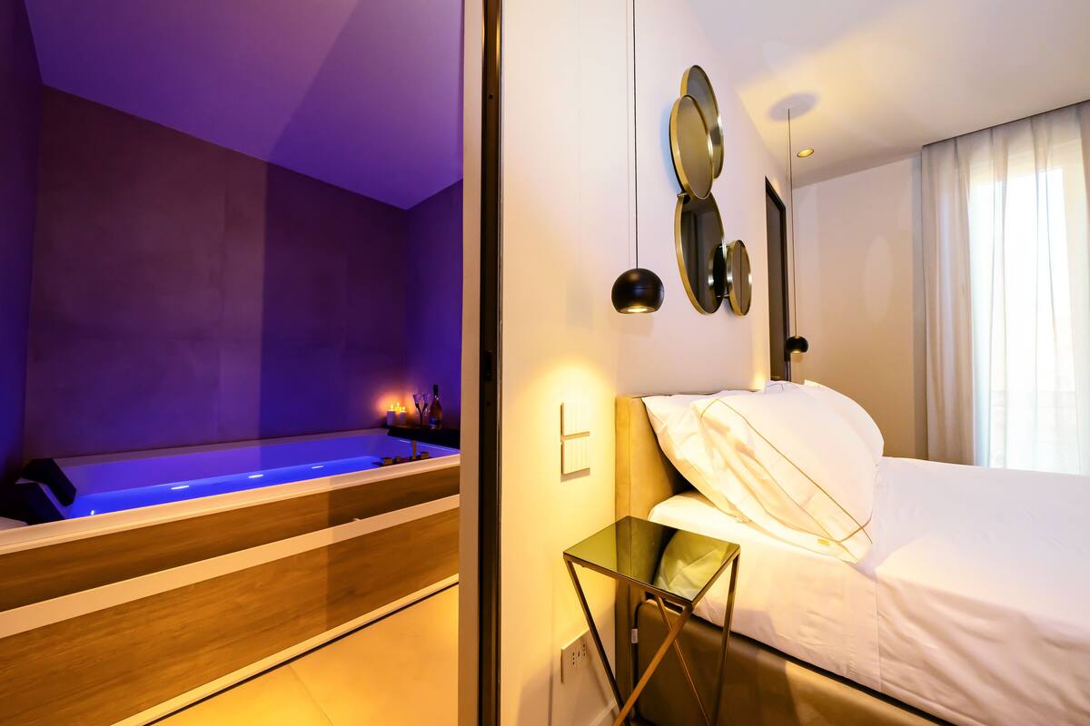 Quarto de hotel moderno com uma cama grande, uma mesa de apoio de vidro, um espelho fixado na parede e uma banheira embutida com iluminação azul.