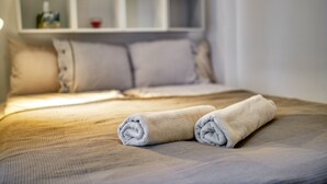 Una cama bien hecha con toallas enrolladas, un cabecero blanco y una estantería con una botella y una taza.