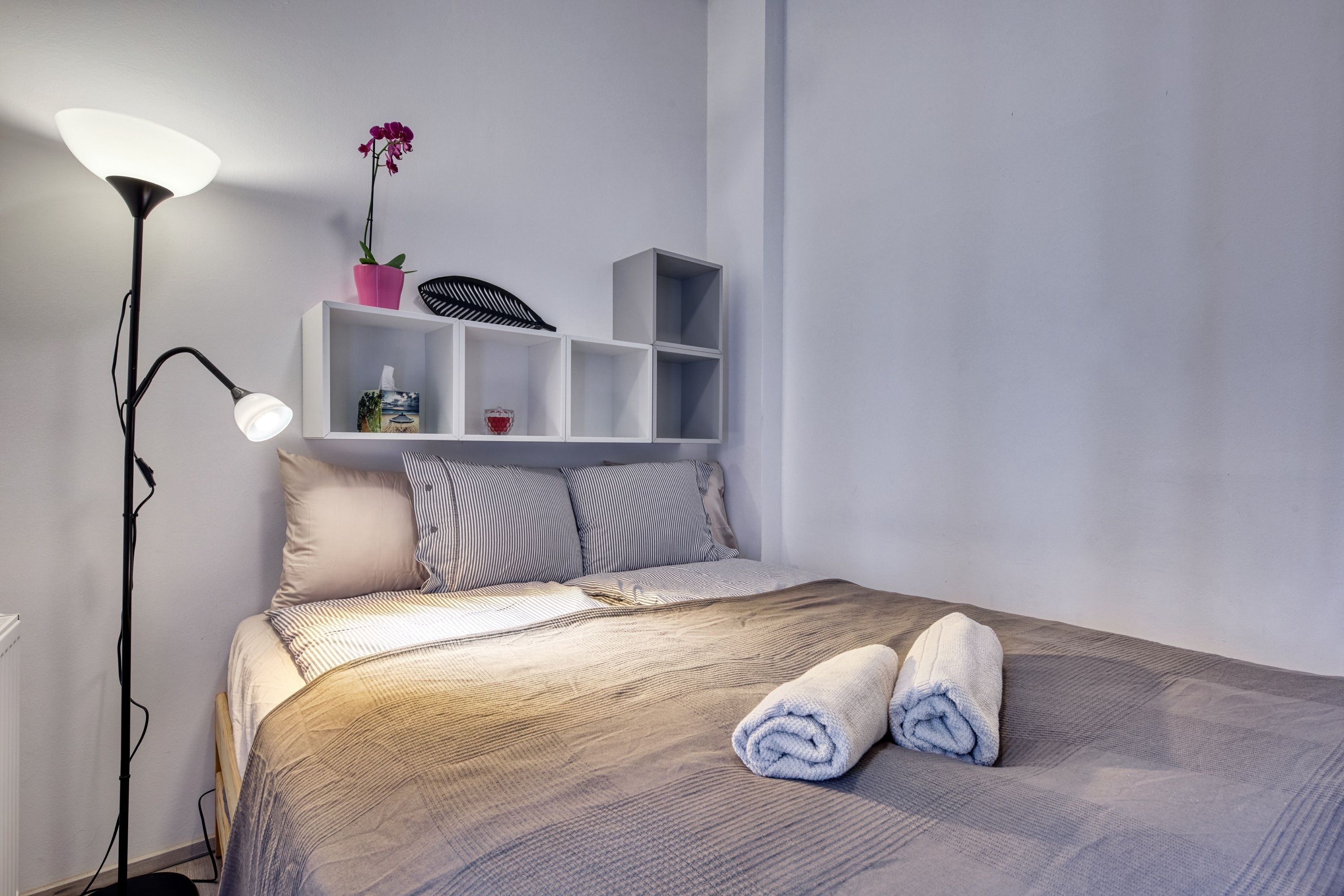 A neatly made bed with a striped blanket, a bedside lamp, and a shelf with a potted orchid and a small box.
