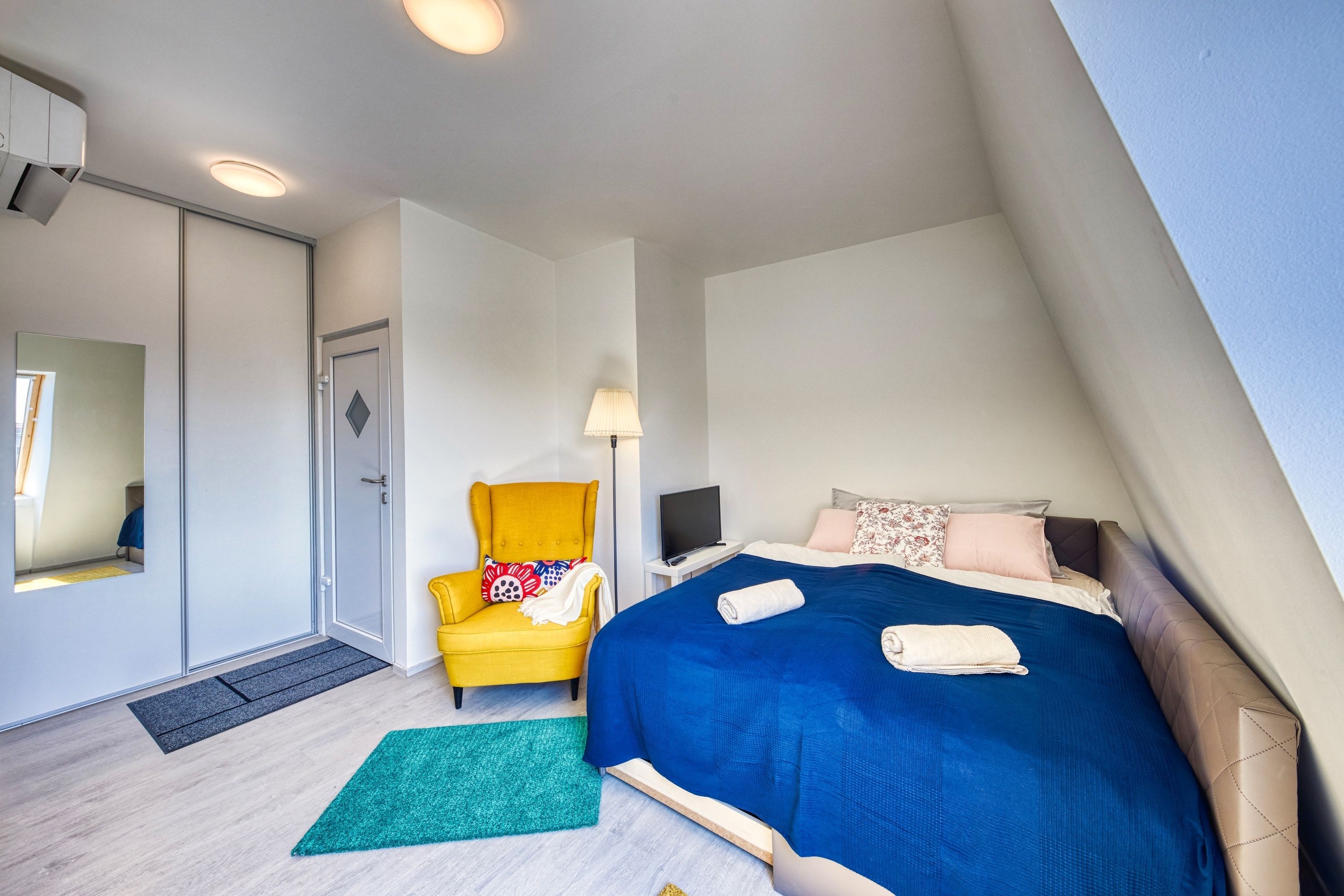 Un dormitorio con cama, una silla, un escritorio con lámpara y un televisor.