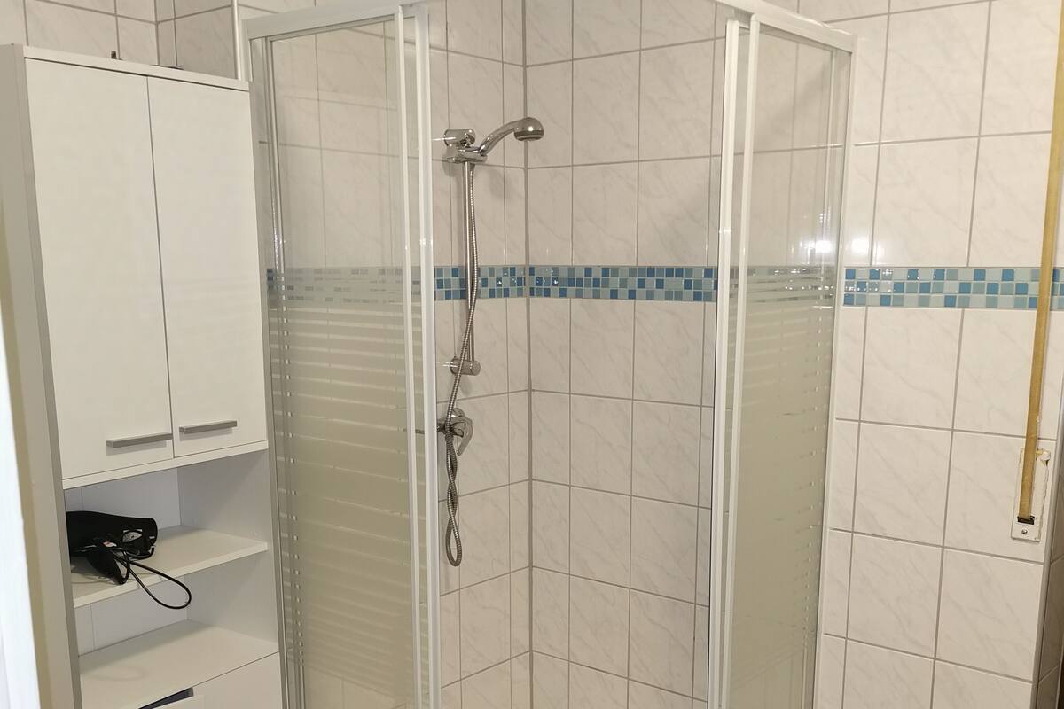 A shower with a glass door, white tiled walls, and a small shelf with a bag.