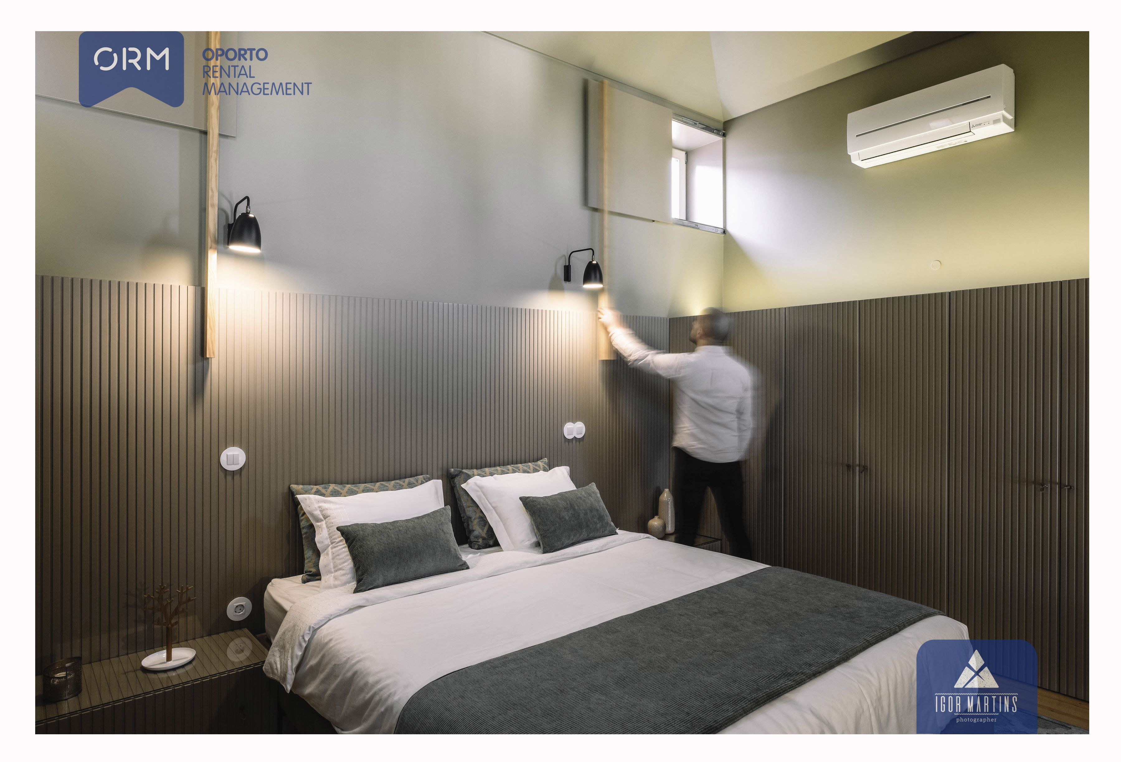 A modern hotel room with a bed, bedside lamps, an air conditioning unit, and a mirror on the wall.