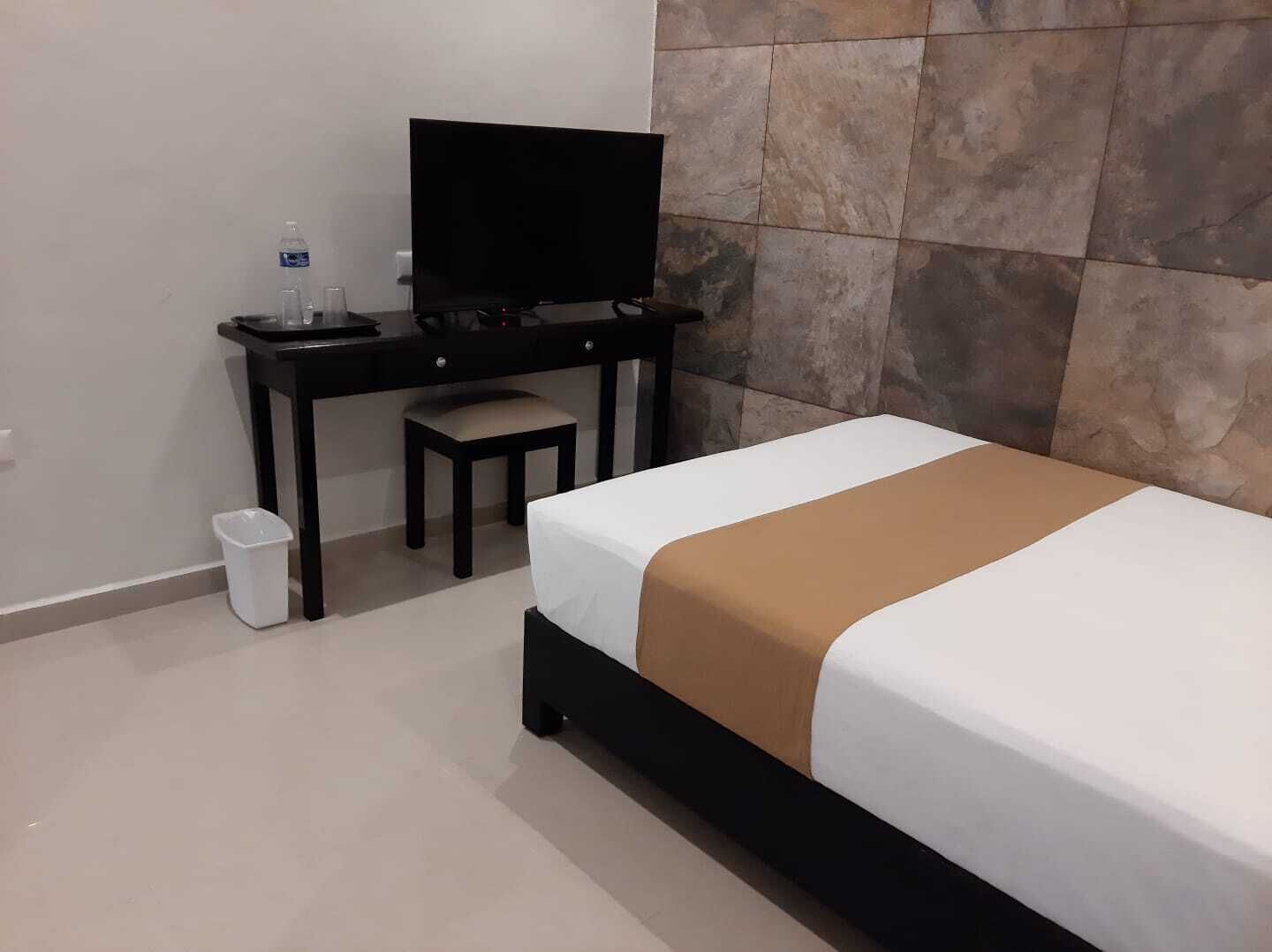 A hotel room with a bed, a desk with a television, a chair, and a trash bin.
