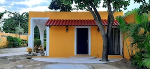 A yellow building with a red-tiled roof, a white door, and a small porch area. - Departamentos X Kanlol (Izamal)