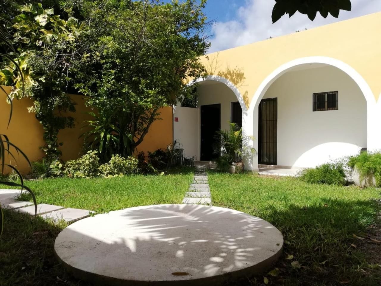 A courtyard with a circular concrete feature, a pathway leading to a building with a yellow facade and white arches, and greenery around.