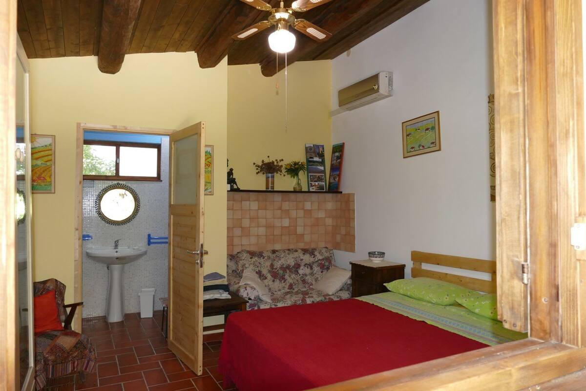 Une chambre avec un plafond en bois, un lit recouvert d’une couverture rouge, une table de chevet en bois, une petite table avec un bol, et une étagère murale ornée de photos et d’un vase.