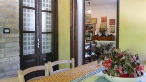 A dining area with a table set for two, a bowl of flowers, and a view into a kitchen area with a hanging light fixture.