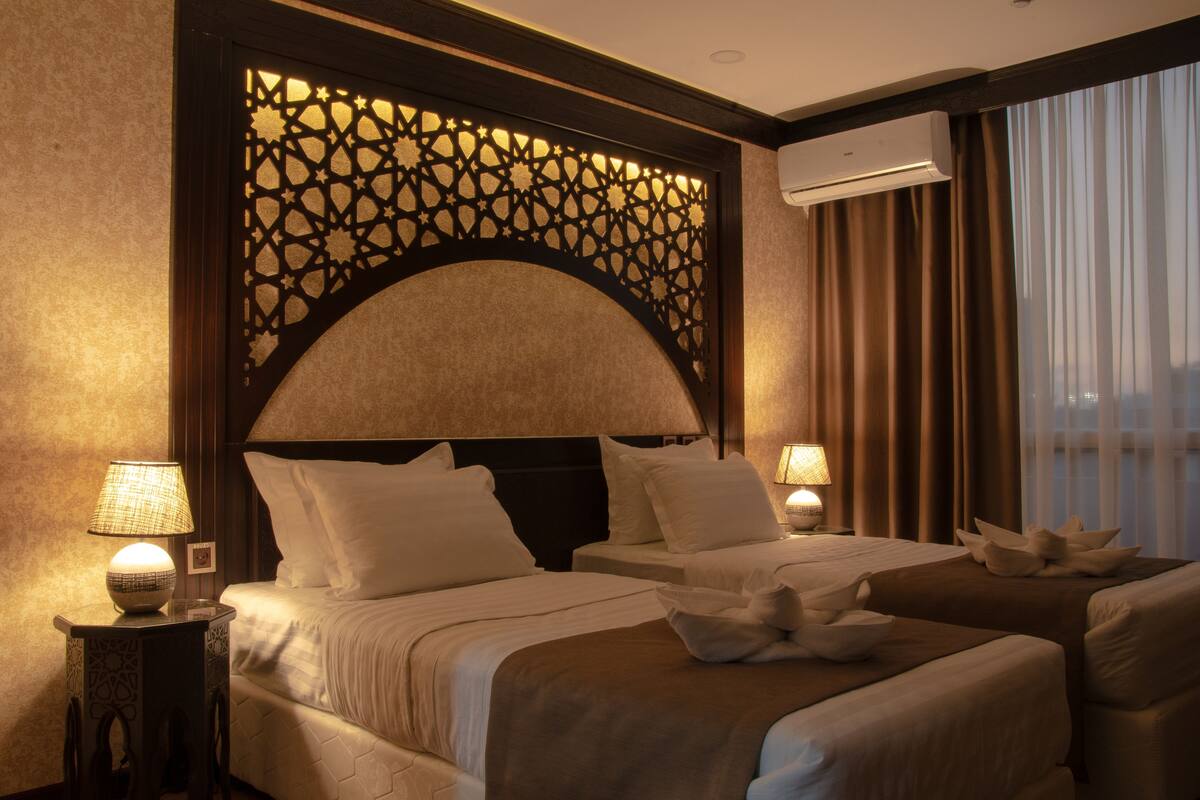 A hotel room with two beds, a decorative headboard, bedside lamps, and a view of the city through large windows.