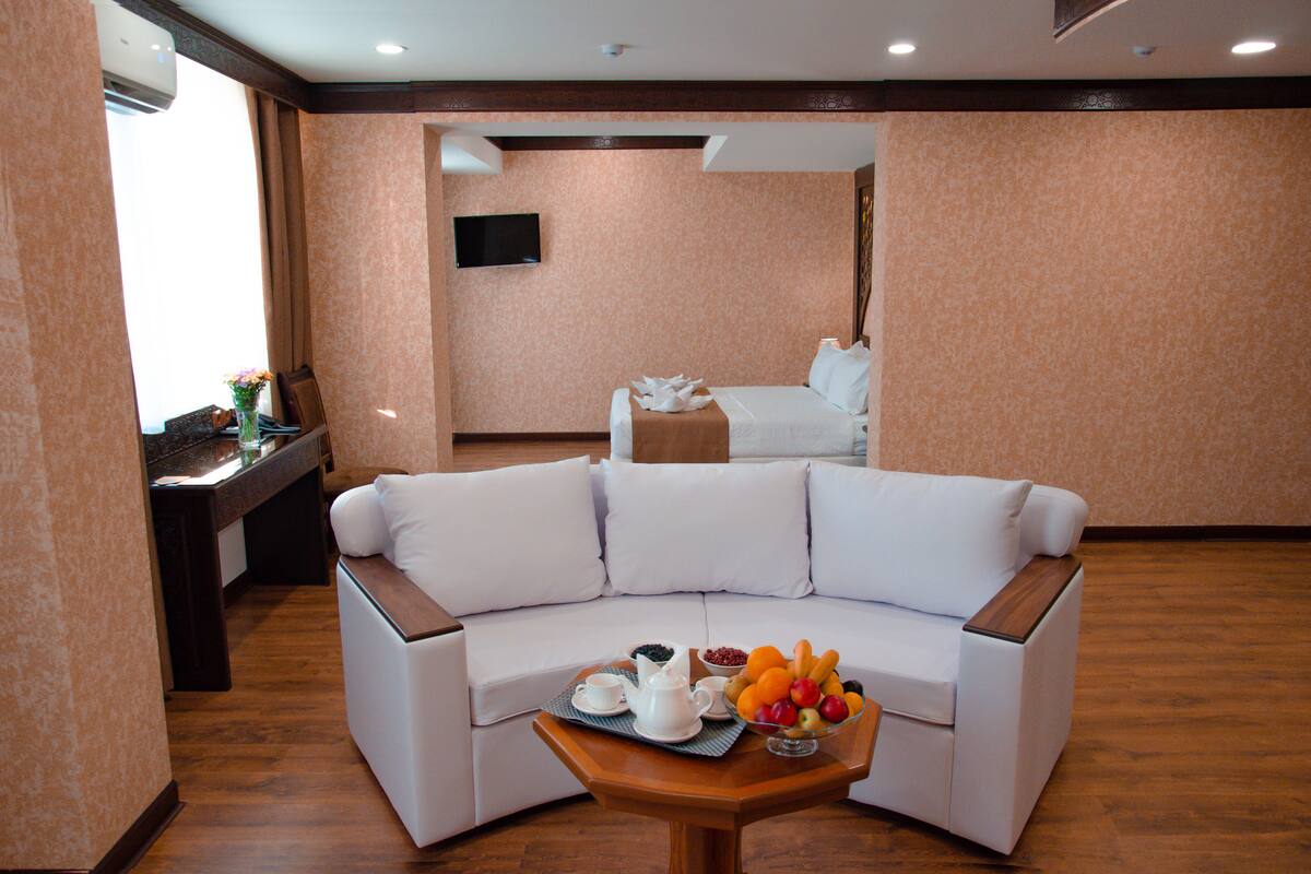 A hotel room with a white sofa, a wooden coffee table with fruits and a teapot, and a bed in the background.