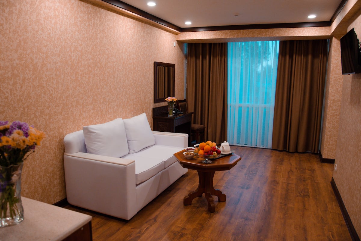 A hotel room with a white sofa, a wooden coffee table with fruits and a teapot, and a vase with flowers.