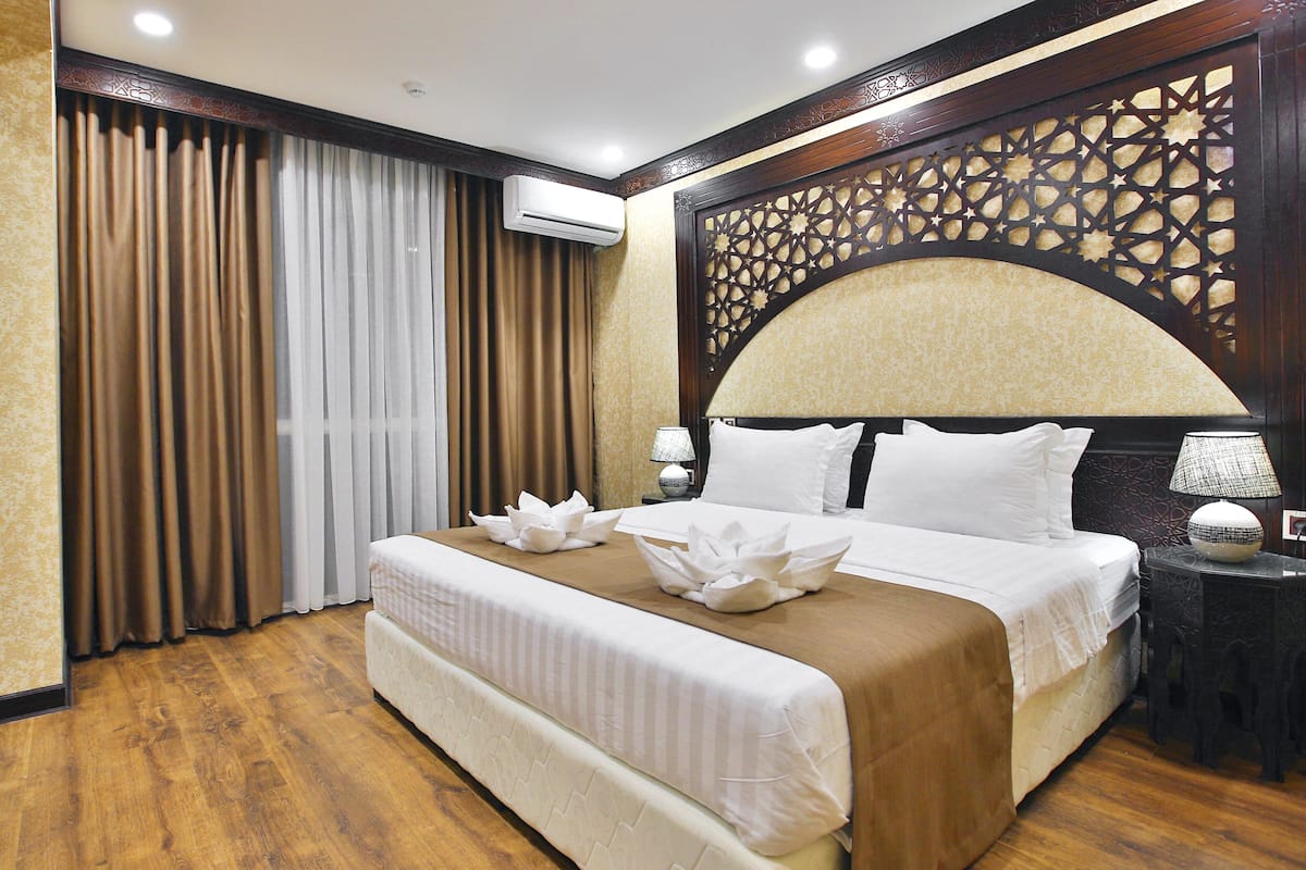 A hotel room with a large bed, a decorative headboard, a nightstand with a lamp, and a window with curtains.