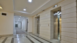 A long, well-lit corridor with a marble floor, two elevators, and a sign indicating the direction to the elevator. - Orient Star Varaxsha (Bukhara)