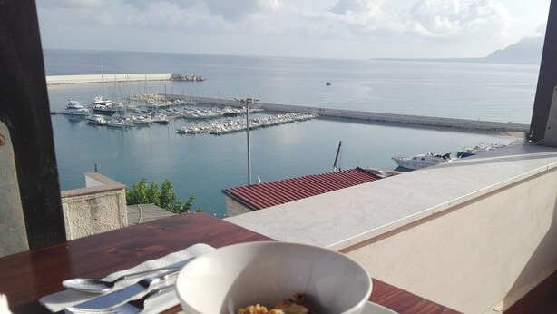 A table with a bowl of cereal and cherries, overlooking a marina with boats and a distant shoreline. - Orlando Bay View (Balestrate)