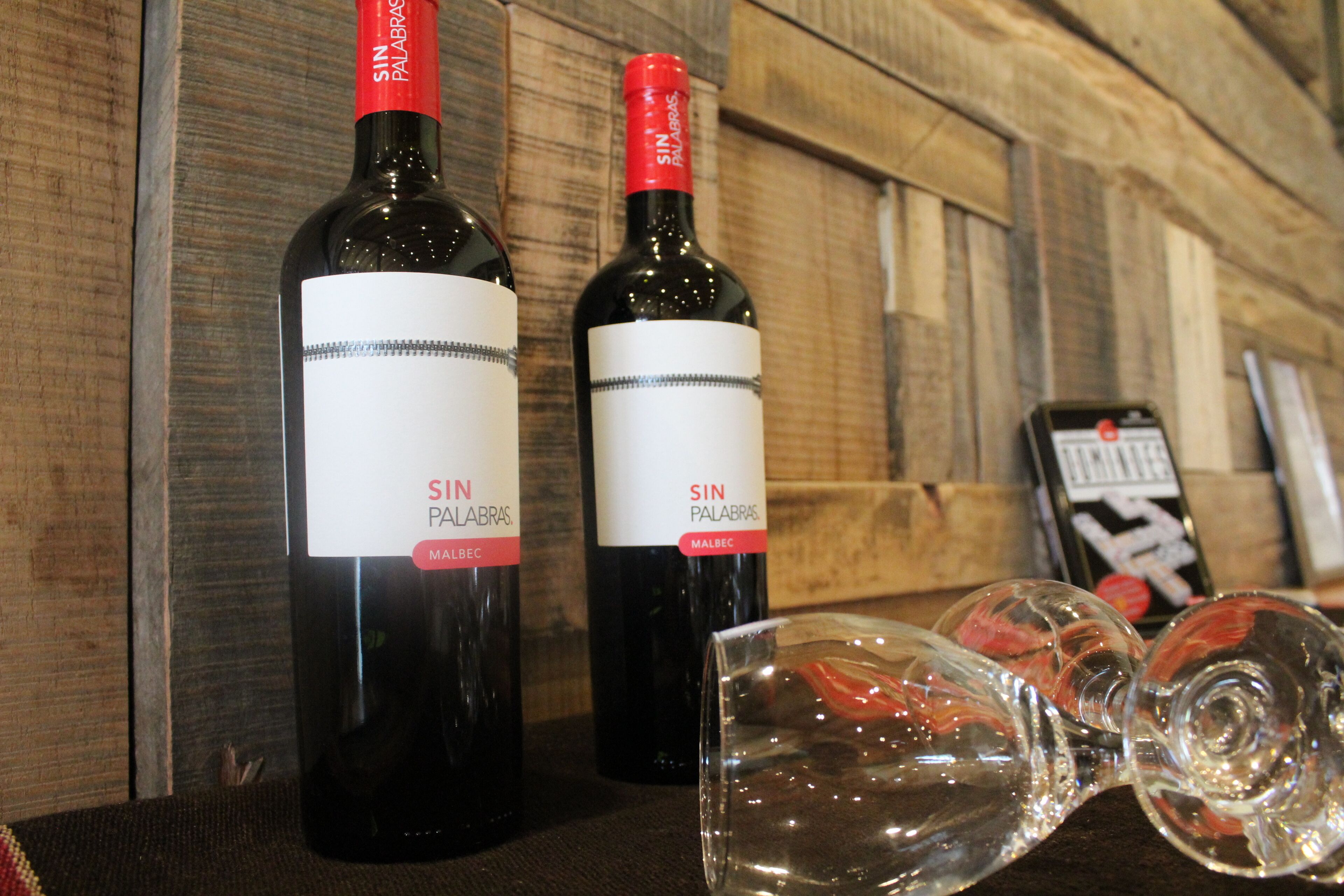 Two bottles of wine with labels, a glass, and a smartphone on a wooden surface.