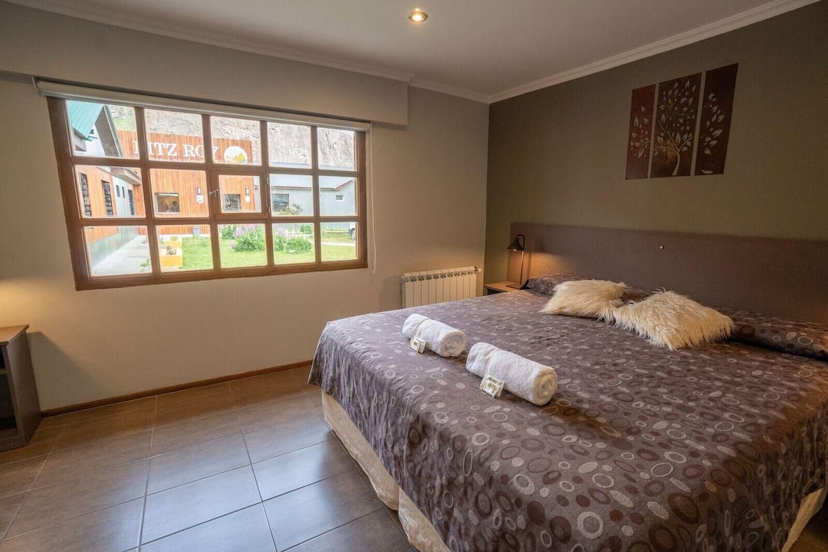 Une chambre avec une grande fenêtre, un lit recouvert d’une couvre-lit à motifs et un radiateur.