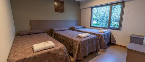 Una habitación con tres camas, cada una con una colcha estampada y una toalla sobre la cama. Se ve una ventana con vistas a árboles.