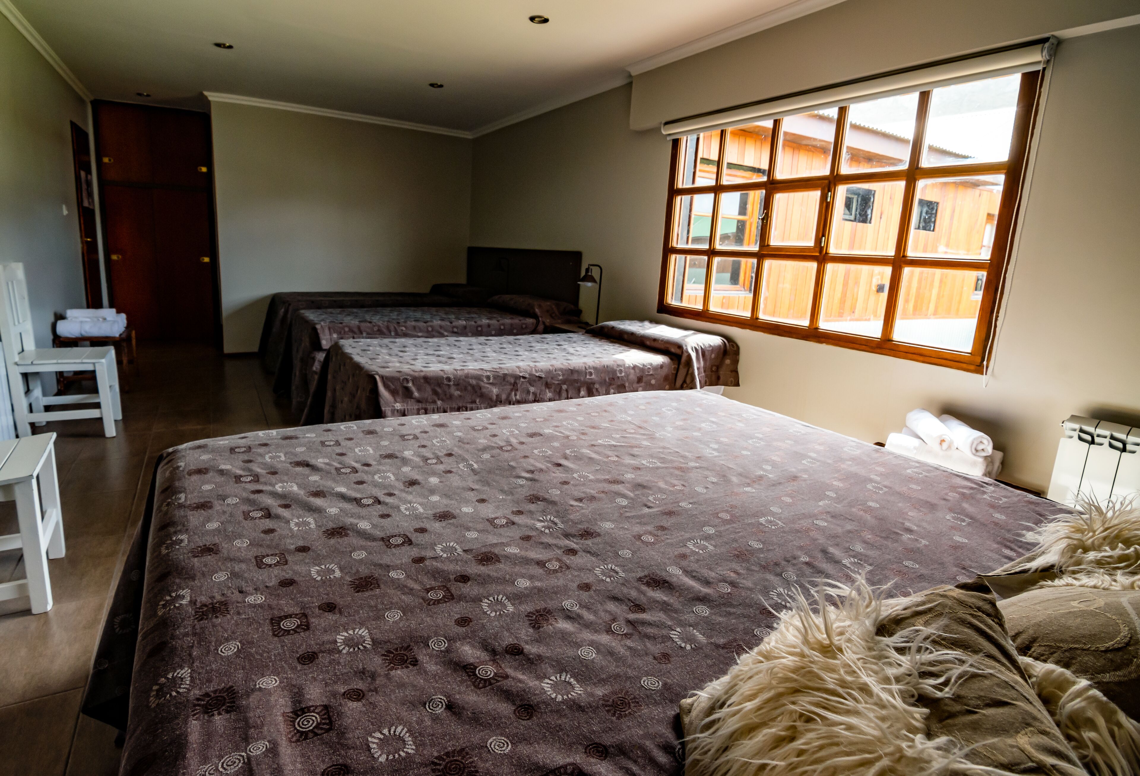 A hotel room with two beds, a desk, and a window with a view of wooden buildings.