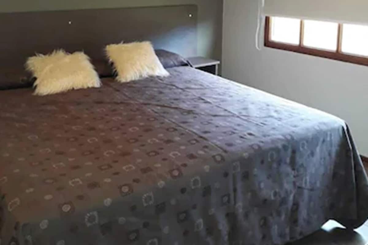 Une chambre à coucher avec un grand lit, deux oreillers, une tête de lit, une table de chevet et une fenêtre avec des stores.