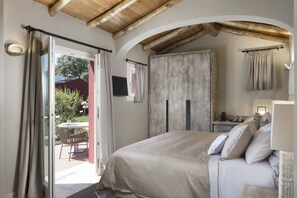 A bedroom with a large bed, a wooden ceiling, and a view of the garden through an open door. - Li Neuli Country Club (Arzachena)