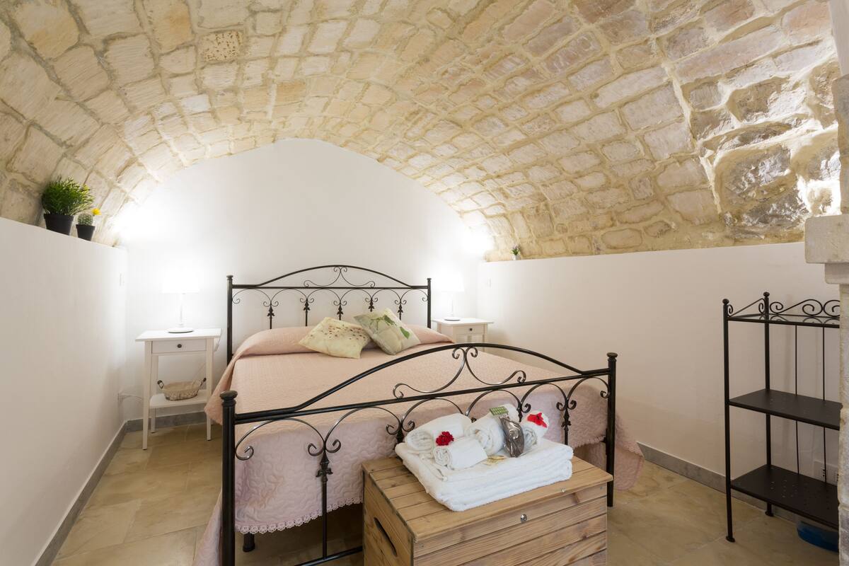 Una camera da letto con soffitto in pietra, un letto con struttura in metallo, un comodino e un settimino in legno.