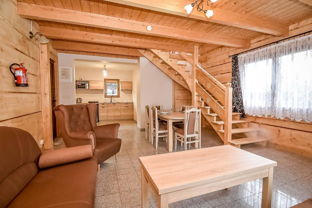 A cozy wooden cabin interior with a sofa, dining table, and staircase leading to a kitchen area.