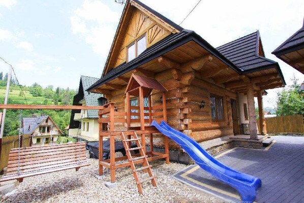 A wooden cabin with a wooden deck, a blue slide, and a wooden play structure.