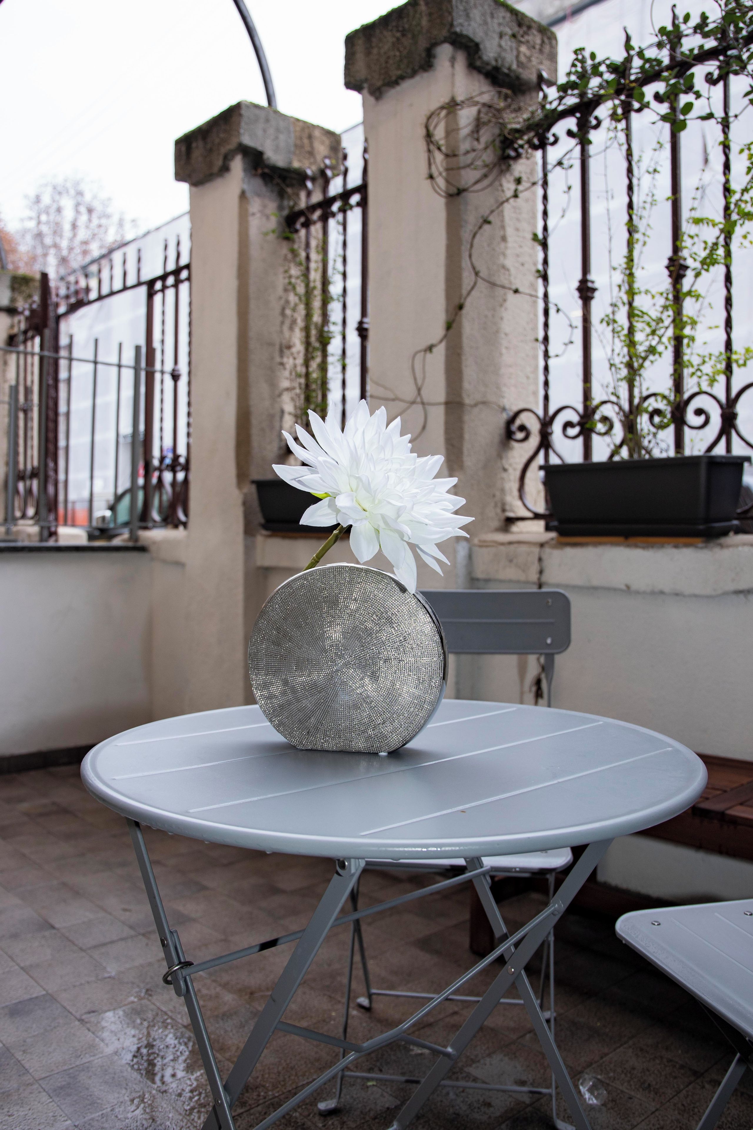 Ein runder Metalltisch mit einer weißen Blumenvase, platziert auf einer gefliesten Terrasse mit einem Metallstuhl.