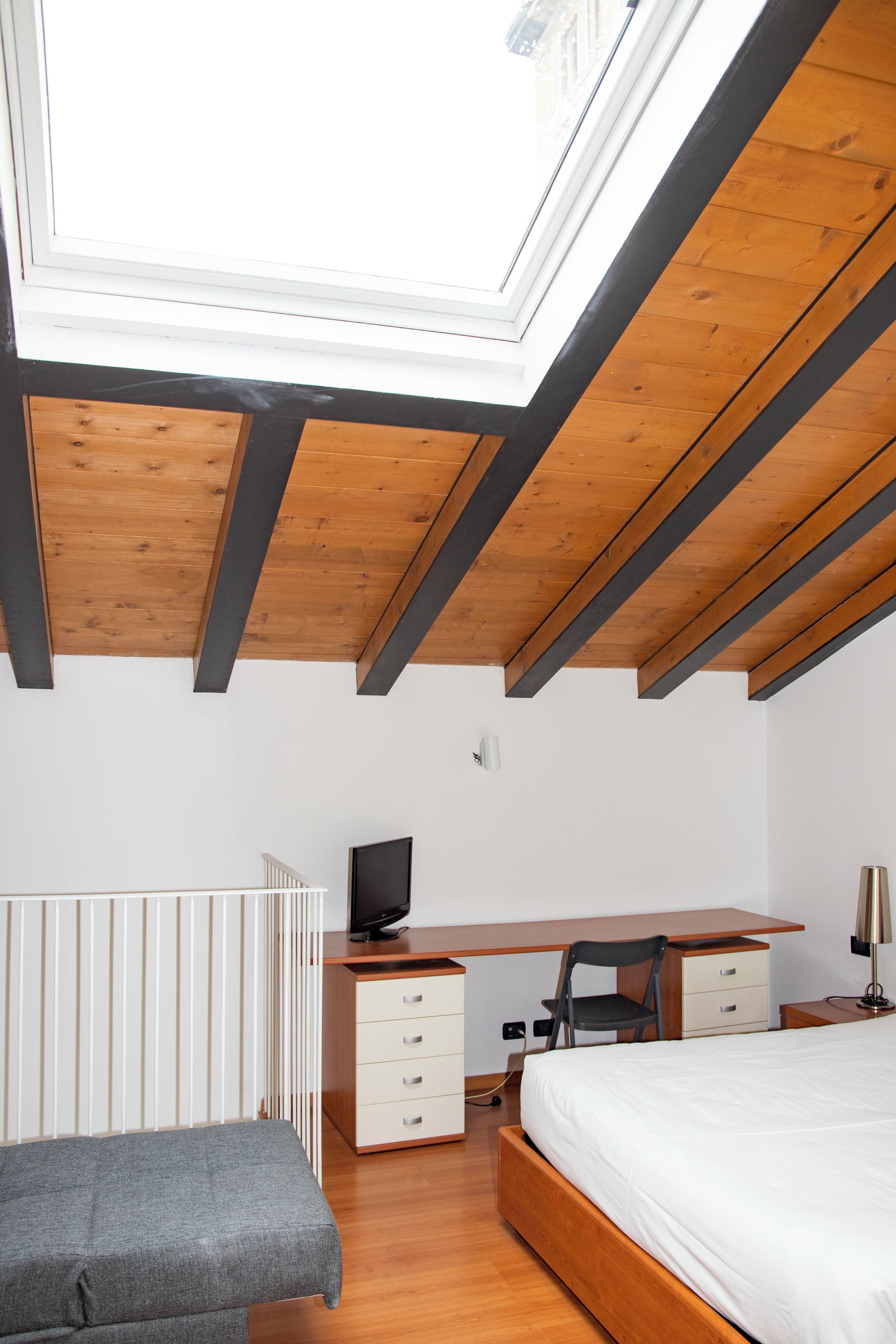 A bedroom with a skylight, wooden beams, a bed, a desk, a chair, and a television.