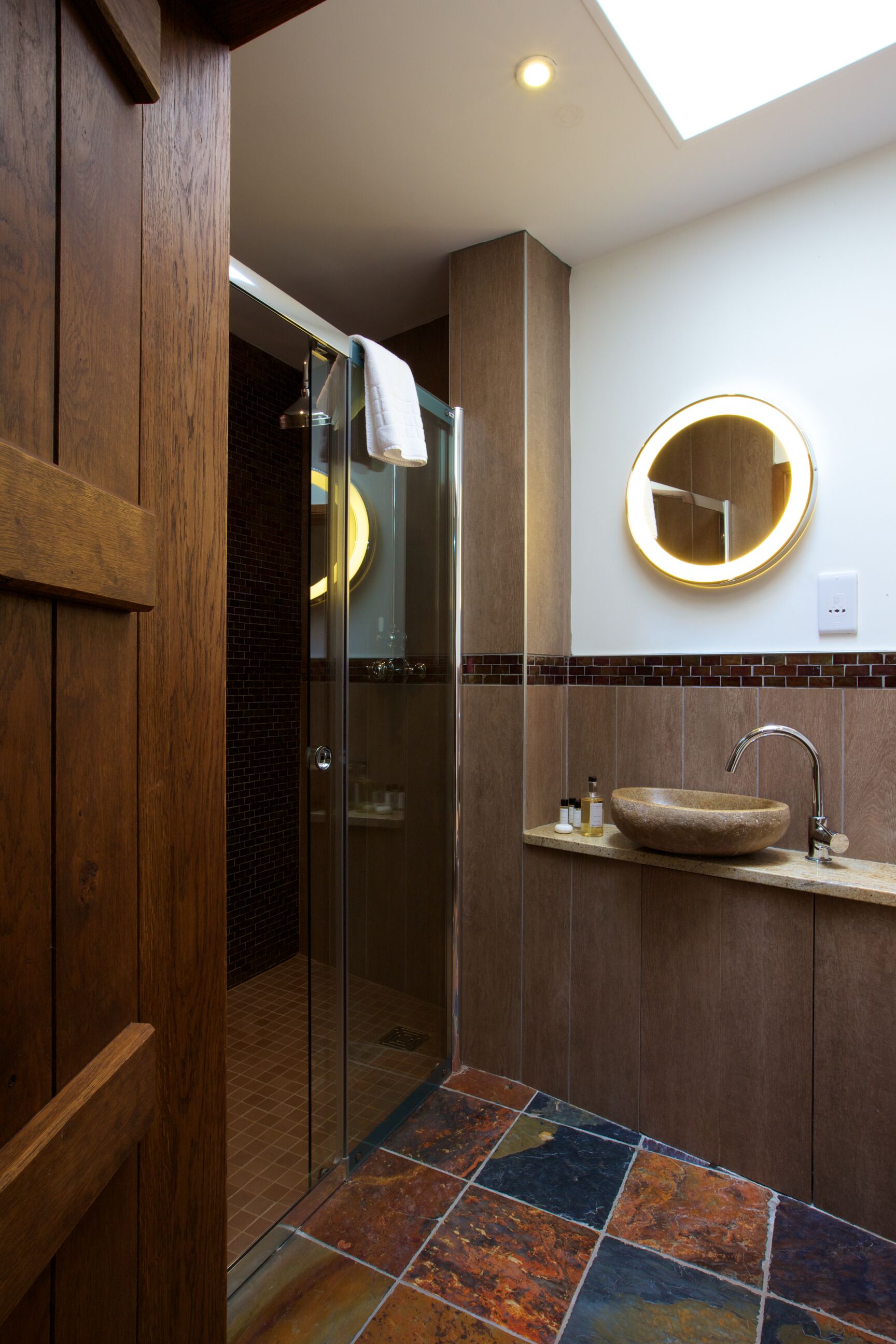 A modern bathroom with a glass shower enclosure, a wooden door, a round mirror with a light, and a stone sink.